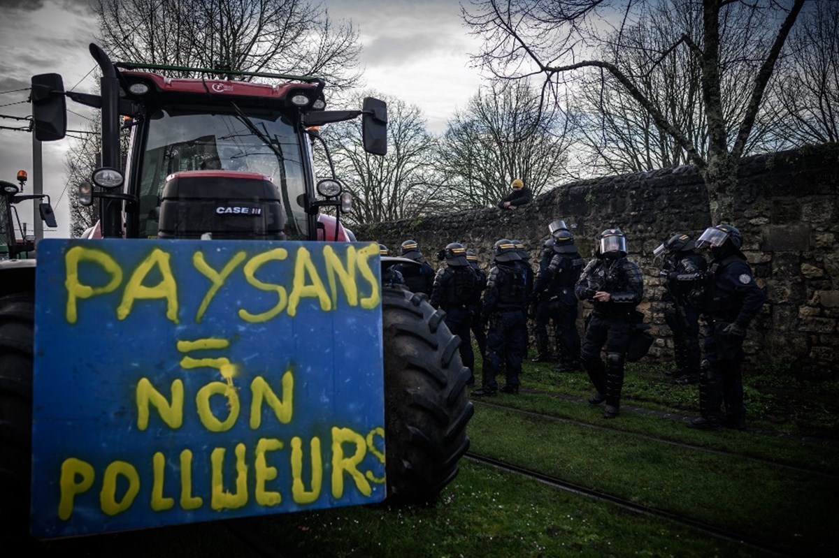 Duvara yaslanan bir çiftçi, "Çiftçiler = çevreyi kirletmez" yazan bir pankart taşıyan traktörün yanındaki polis memurlarına bakıyor.